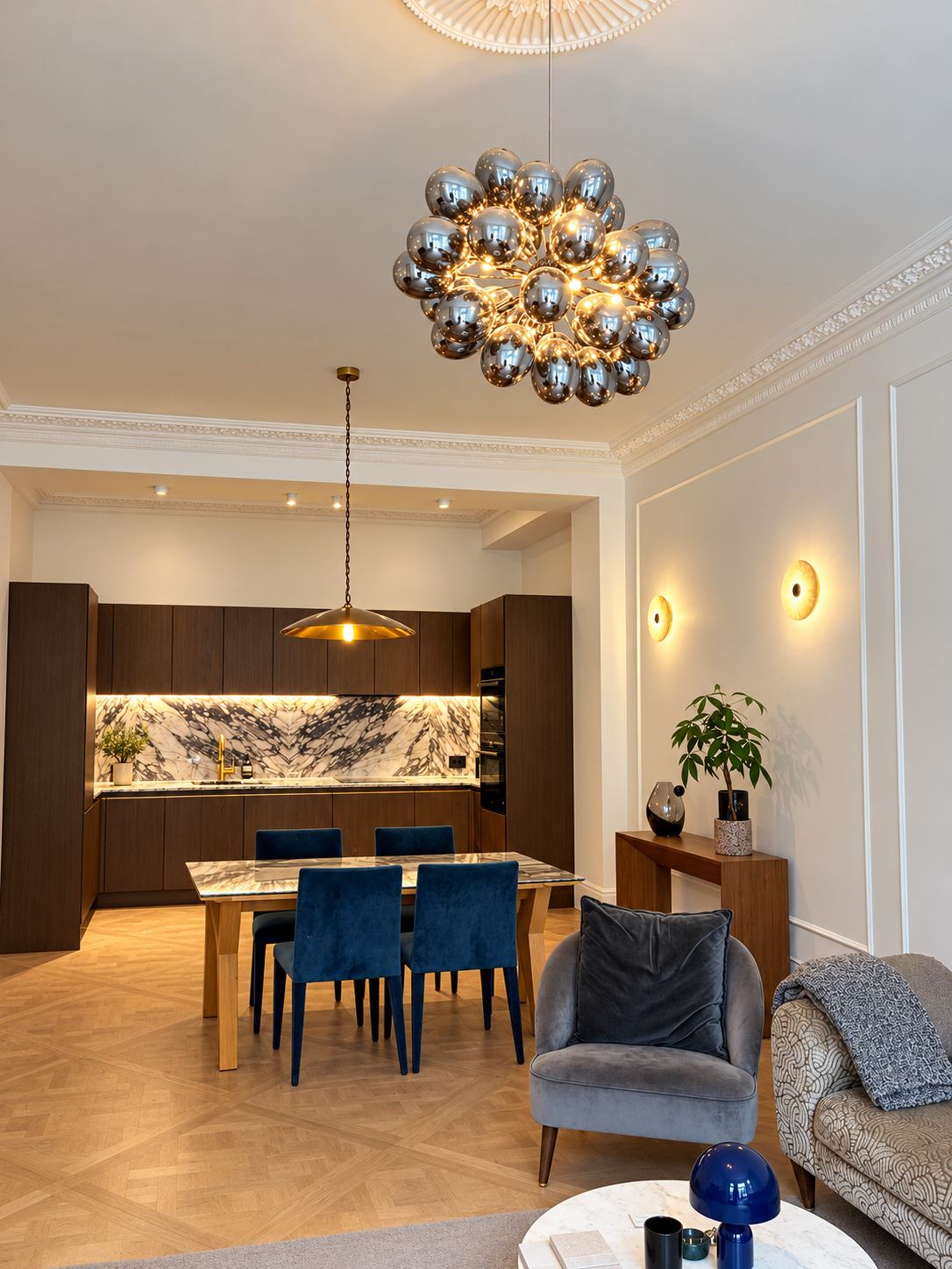 Open-plan living, dining and kitchen area with a chrome cluster chandelier, decorative wall lighting and an illuminated kitchen.