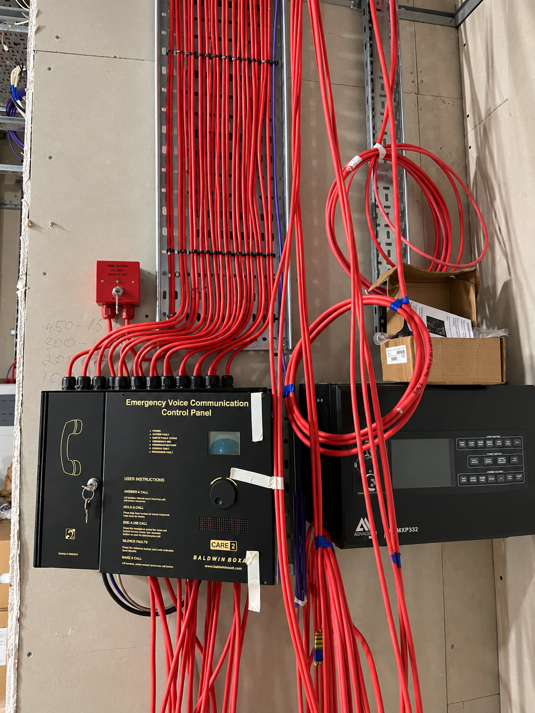 Emergency voice communication panel and a fire safety panel on an unfinished wall with red cables bundled along metal cable trays.