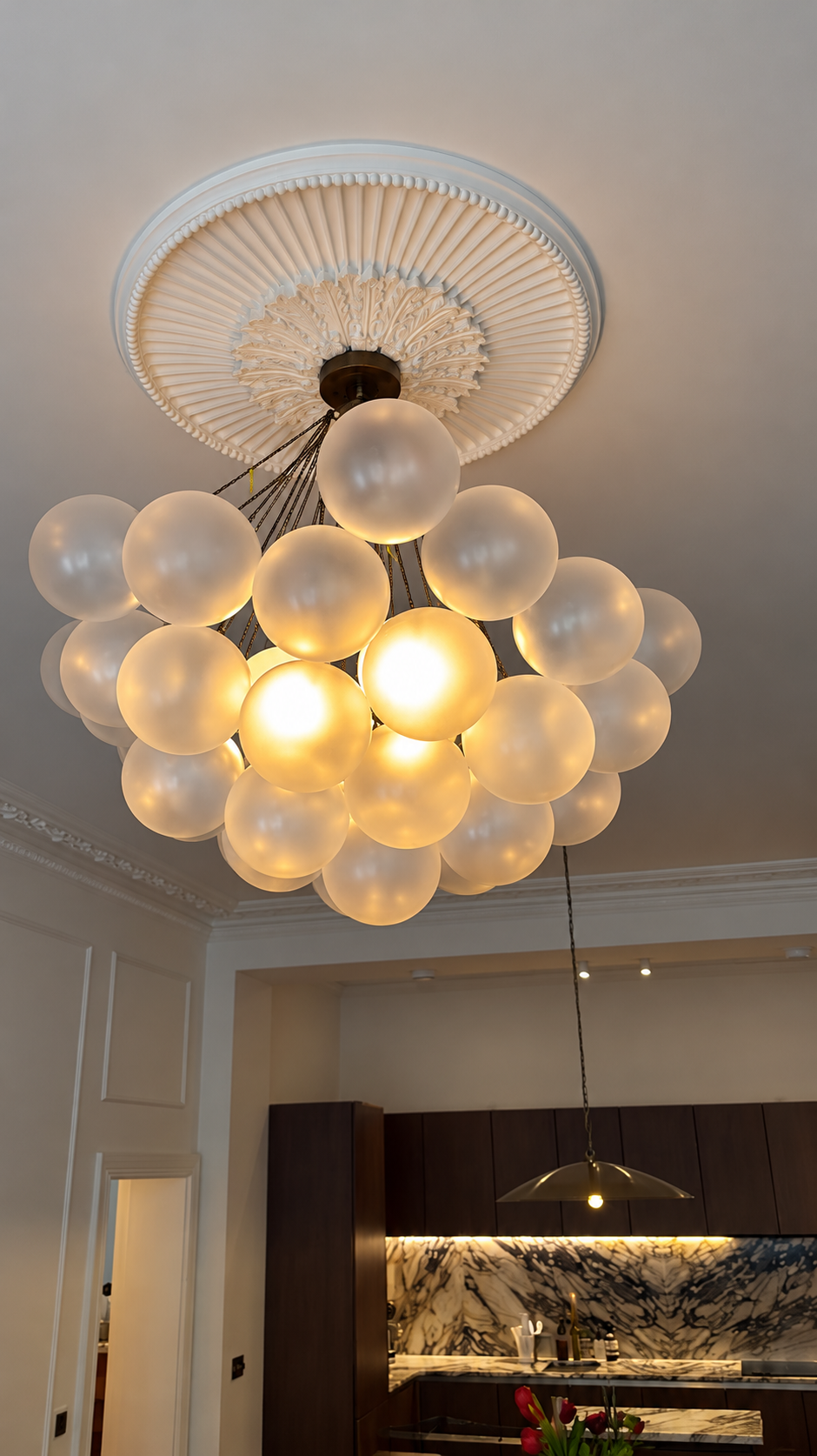 Large chandelier of illuminated frosted glass globes hanging from a ceiling rose above a kitchen with dark cabinets and marble surfaces.