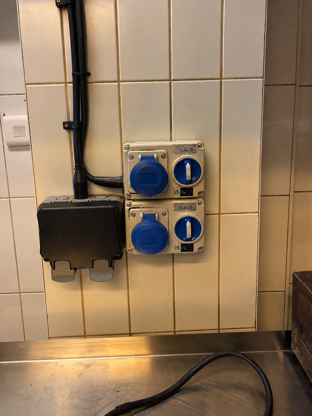 Industrial sockets and an outdoor switch box with surface-mounted conduit on a tiled wall above a stainless steel work surface.