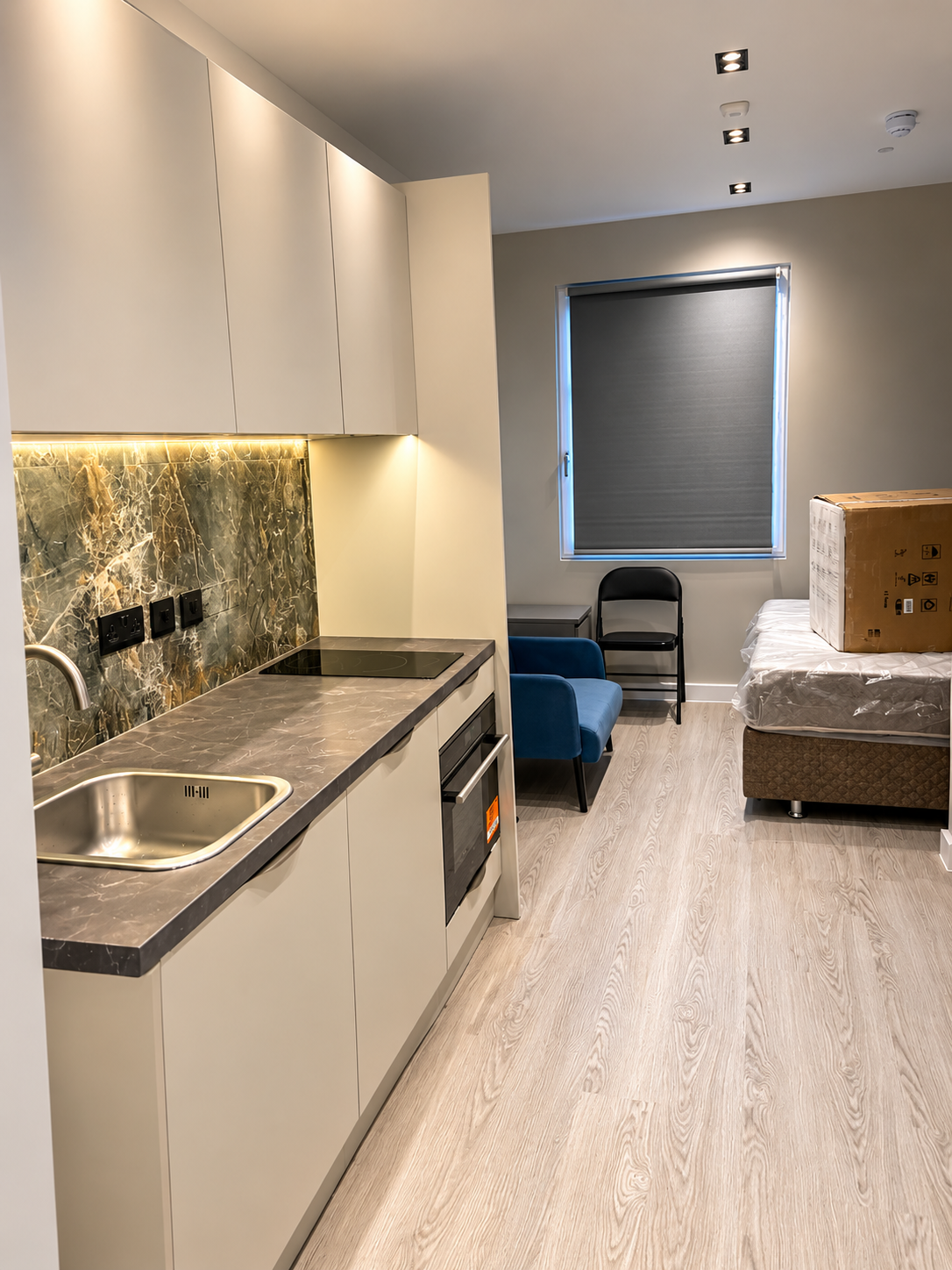 Studio interior with a kitchenette, dark worktops and an illuminated marble-patterned splashback leading to a lounge and sleeping area.