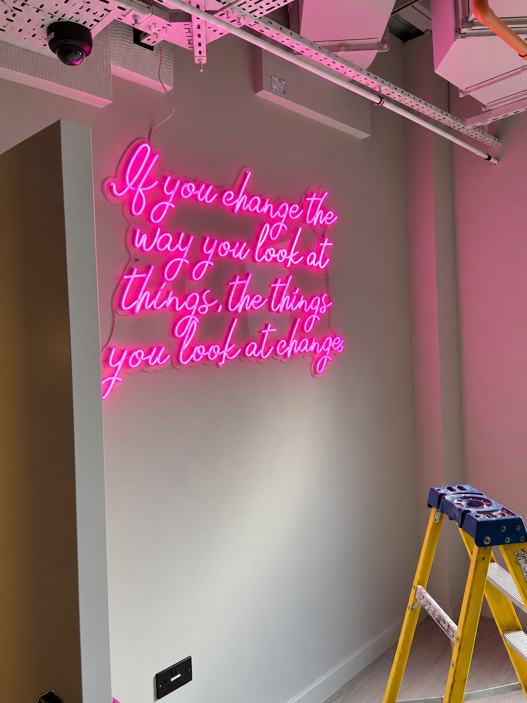 Pink neon sign with cursive lettering mounted on a light grey wall beside an open ceiling.