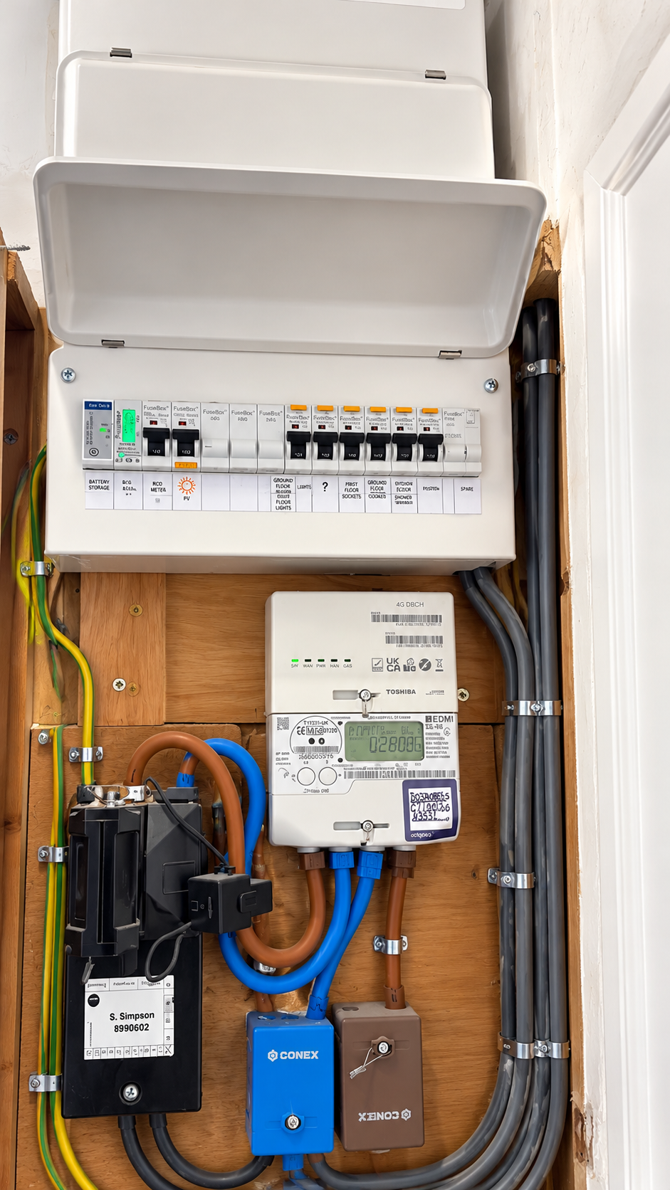 Open white consumer unit mounted above an electricity meter with wiring and connection boxes on a wooden backing board.