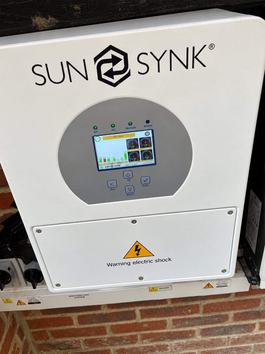 White Sunsynk solar inverter mounted on an exterior brick wall with a digital display, status lights and a shock warning label.