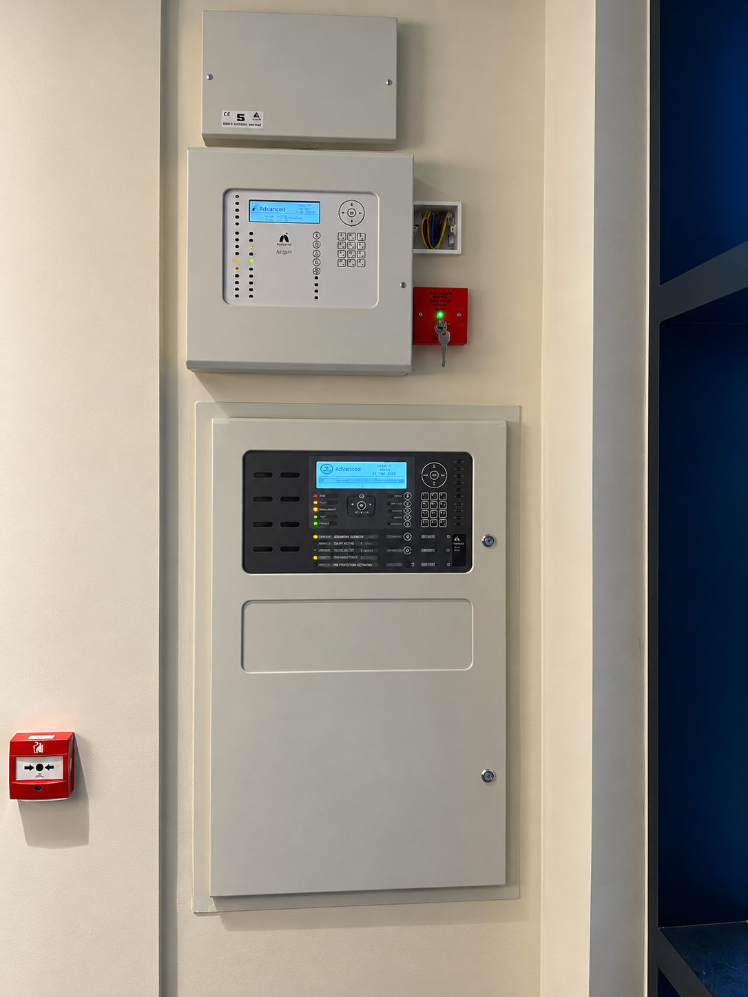 Grey wall-mounted fire alarm control panels, switches and a red manual call point on a light wall.