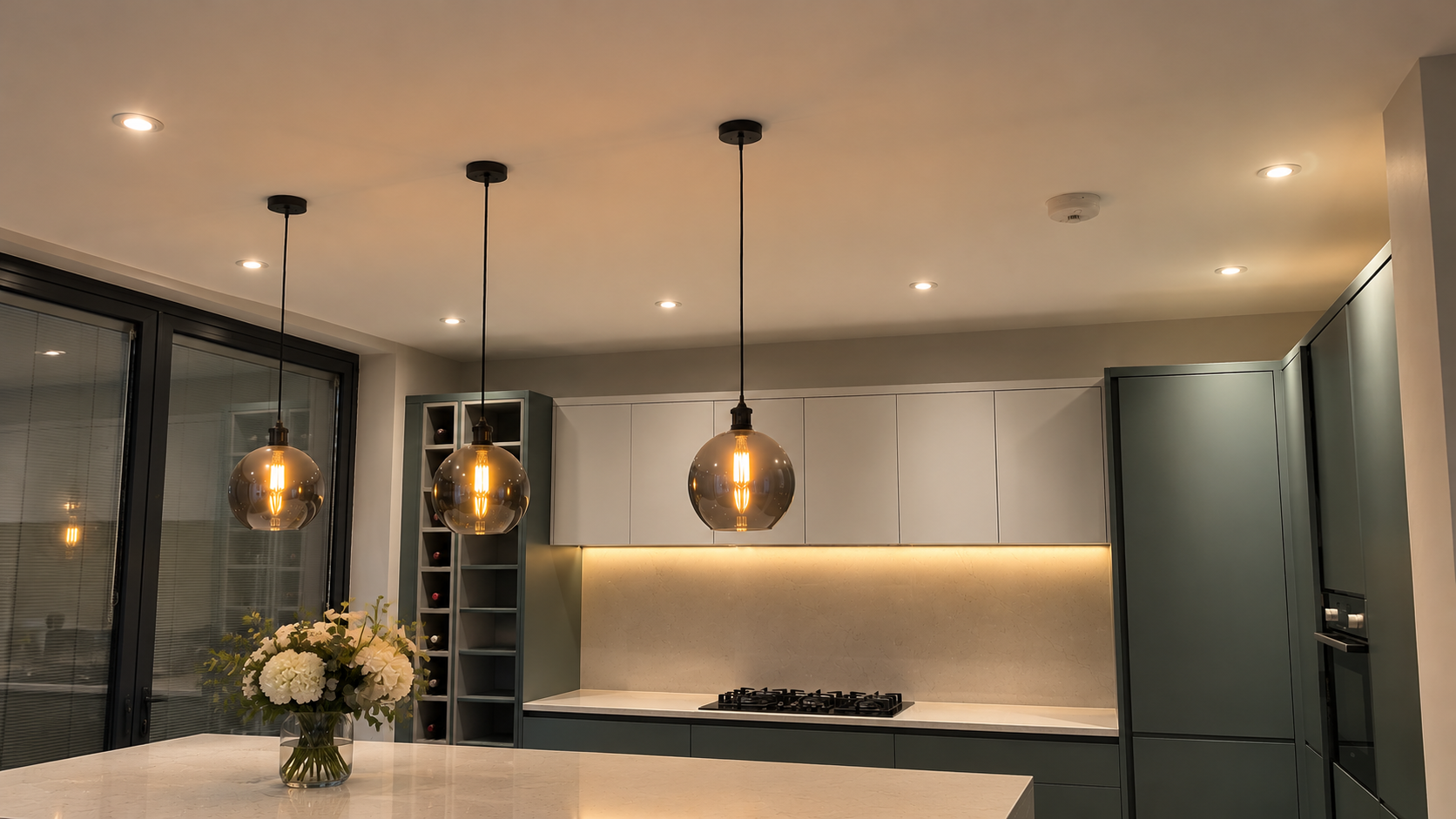 Kitchen with a row of three smoked-glass globe pendant lights over the island, recessed ceiling downlights and warm under-cabinet LED lighting.